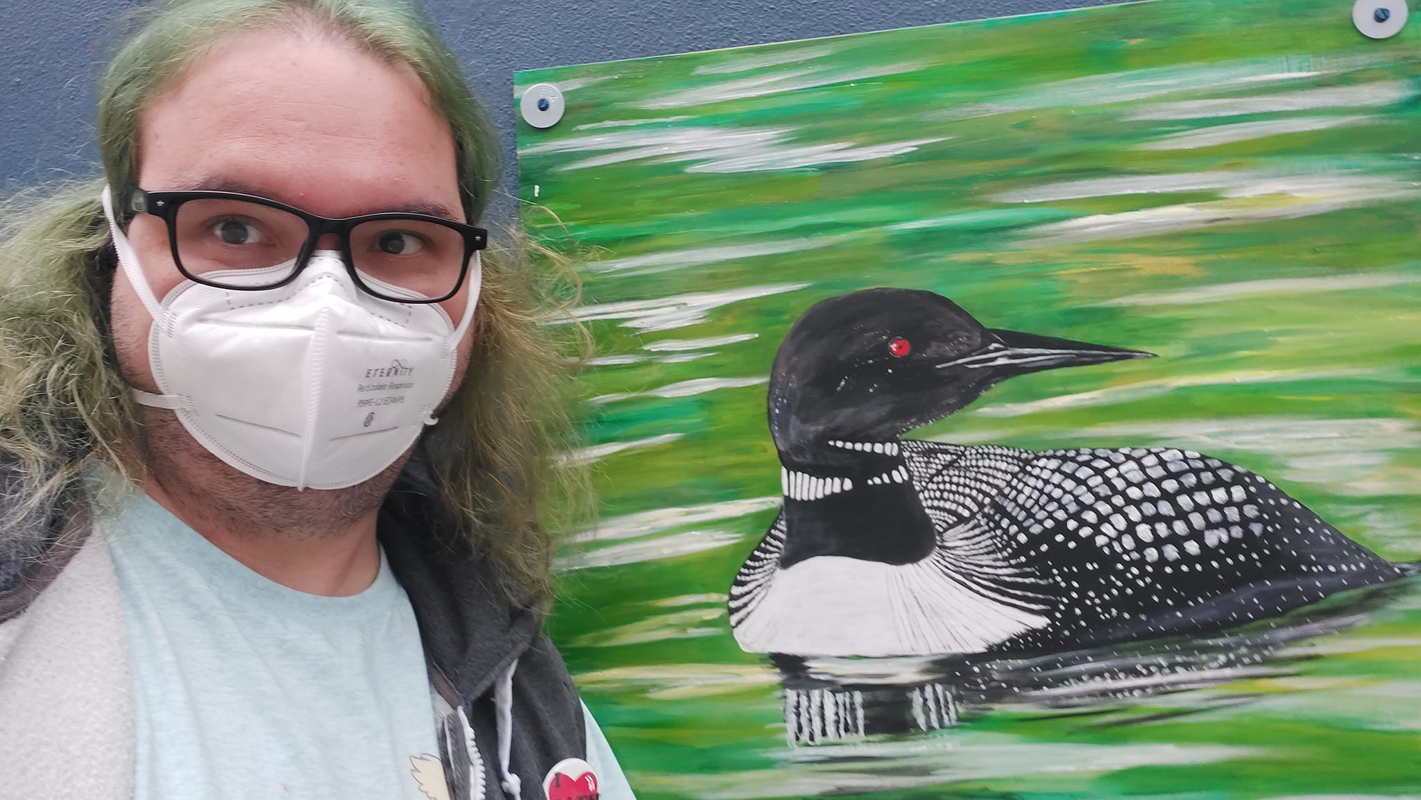 A man with long green hair and no beard wears a mask in front of a painting of a common loon